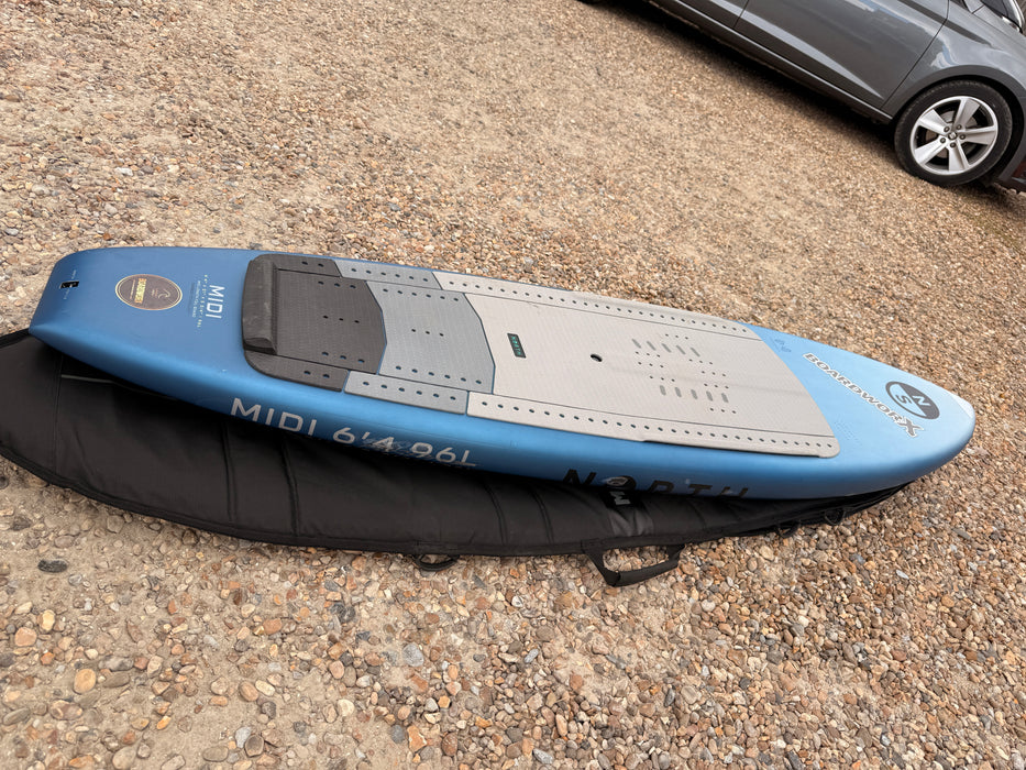 North Midi 6'4 96L wing foil board, side rail profile resting on its Mystic padded board bag