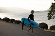 Person in wetsuit carrying blue Duotone Crest D/LAB wing foil board by the seaside