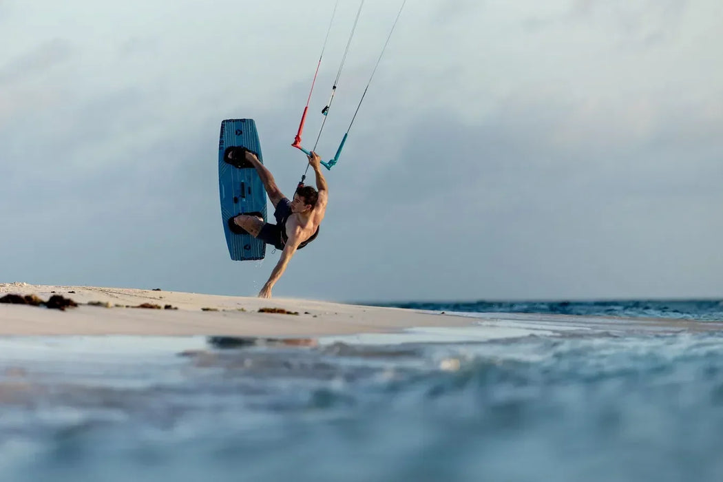 Man kitesurfing with a blue Duotone kiteboard on a beach, performing aerial trick