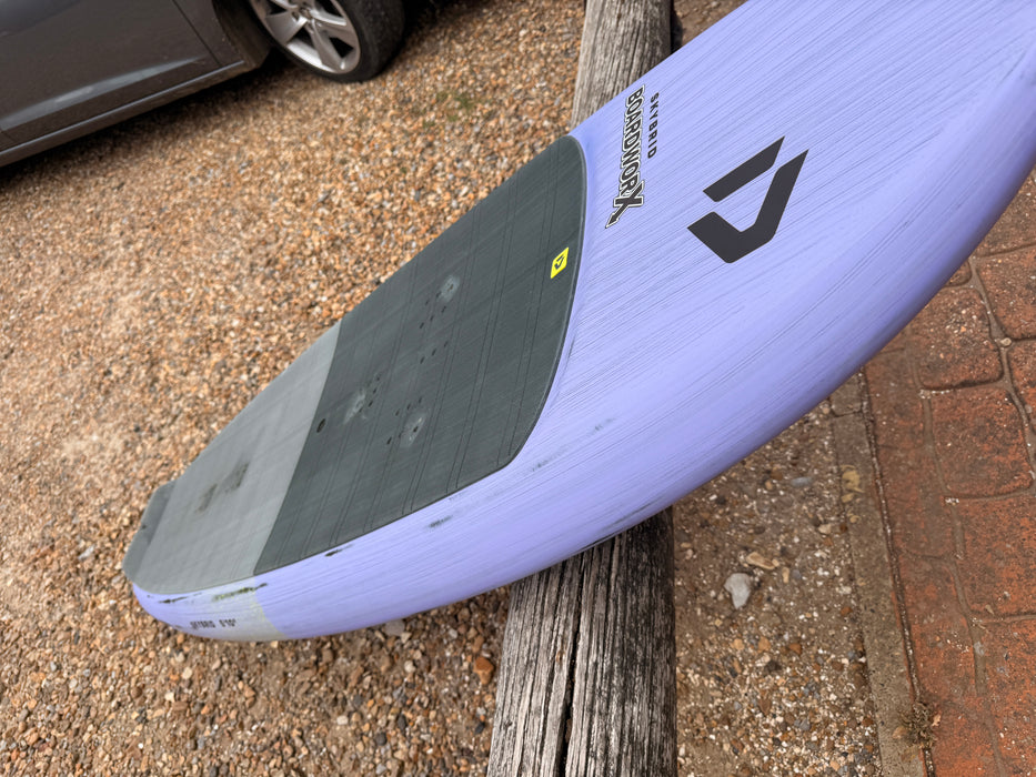 Tail rail profile of used Duotone Skybrid 5'10" 100L 2025 wing foil board leaning against fence showing complete board outline