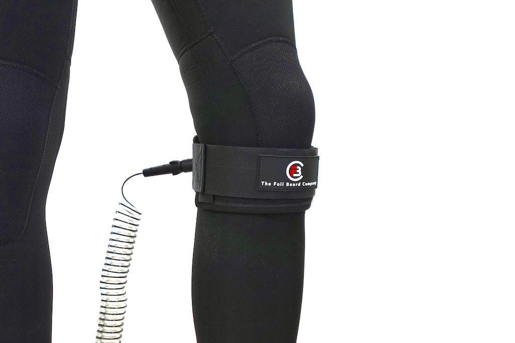 The Foil Board Company coiled wingboard knee leash in black, worn on wetsuit-clad leg.