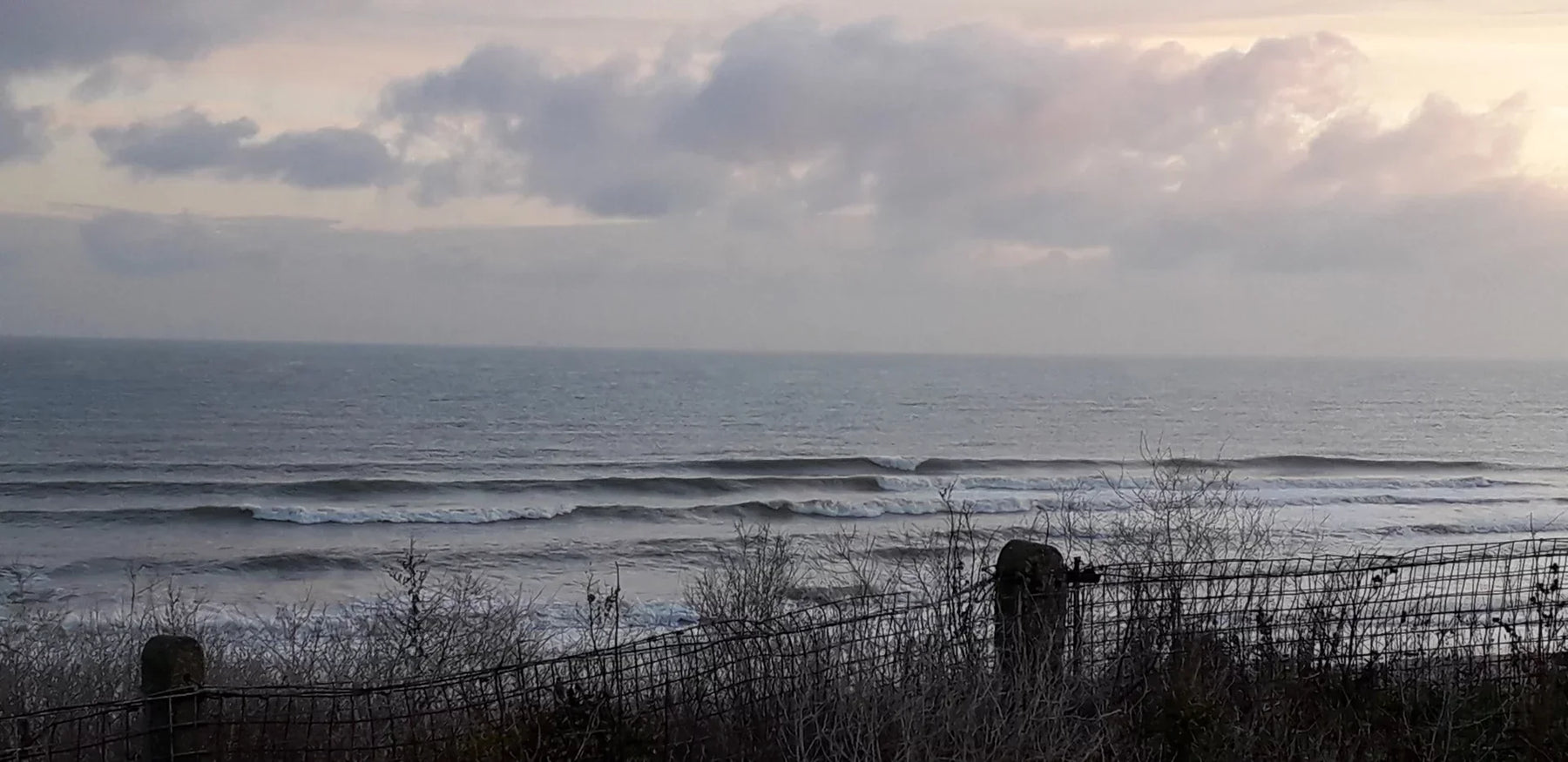 Kent Surf coastline with rolling waves at sunrise, cloudy sky, and seaside fencing visible