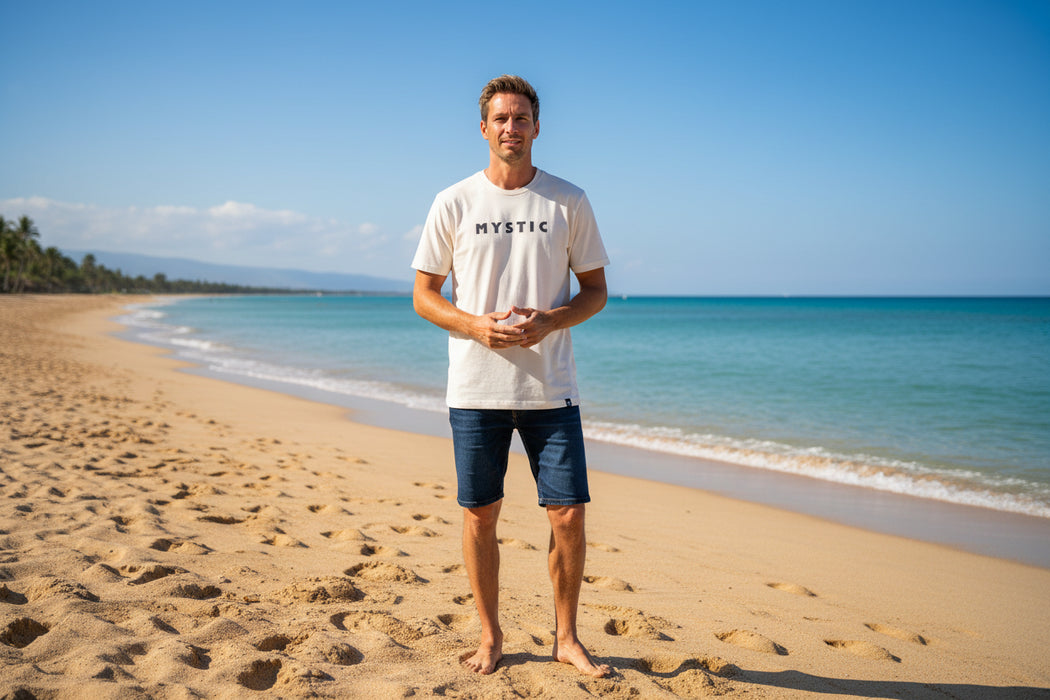 Man wearing off-white Mystic brand t-shirt and denim shorts on sandy beach by the sea