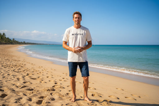 Man wearing off-white Mystic brand t-shirt and denim shorts on sandy beach by the sea