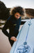 O'Neill black chest zip wetsuit, man waxing O'Neill surfboard on sandy beach