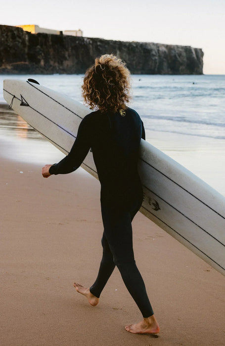 Person in black O'Neill wetsuit carrying long surfboard on sandy beach, ocean and cliffs in background