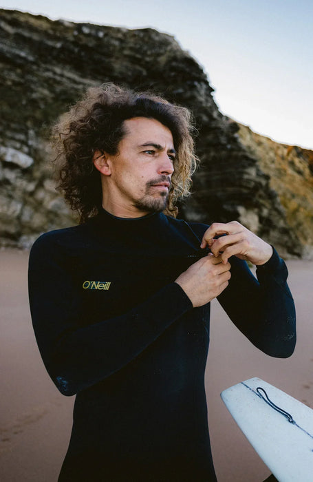 Man wearing black O'Neill chest zip wetsuit on beach, holding a surfboard