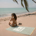 Slowtide Out of Office woven beach towel in green on sand, woman in bikini by the sea
